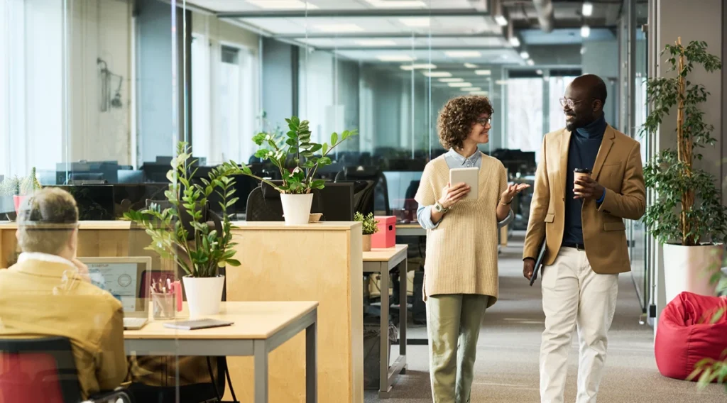 An inviting office space decorated with plants, with two businesspeople chatting with coffees.